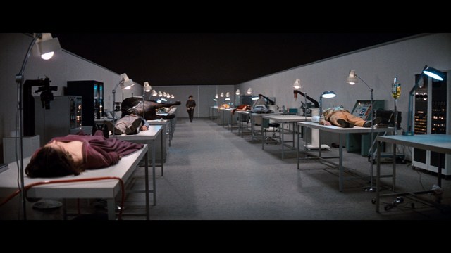 A scene from a movie featuring a man dressed in a bishop's robe speaking at a podium surrounded by microphones, with a woman in a red outfit smiling next to him, and an audience in the background.