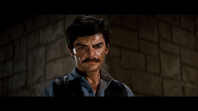 A man with long hair and a baseball cap appears shocked or surprised, sitting in a dimly lit room with wooden paneling and chairs in the background.