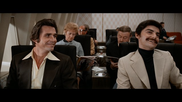 A man with a beard and mustache smiles warmly while standing on a boardwalk by the beach, with colorful flags overhead and a large building in the background.