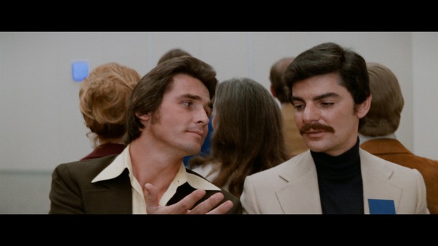 A serious-looking cardinal stands in front of a wooden backdrop with colorful posters related to Catholicism, holding a microphone at a press conference.