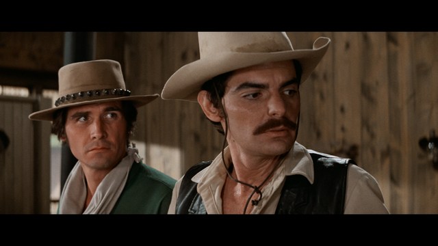 A woman with a pensive expression rests her chin on her hand, sitting at a table with kitchen appliances and items in the background.