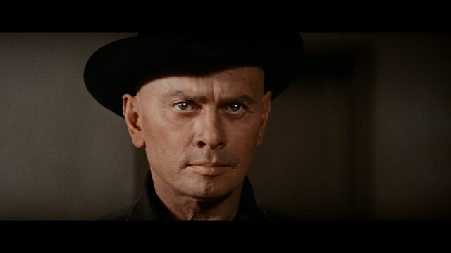 A man in a white suit and hat stands at a doorway, smiling, with a suburban landscape in the background.