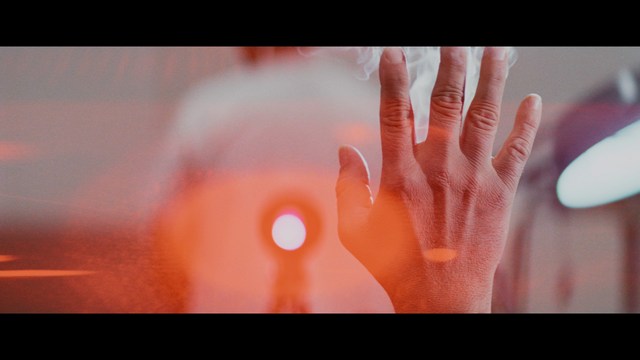 A man with large, feathered angel wings stands in a room, wearing a black suit and looking directly at the viewer.