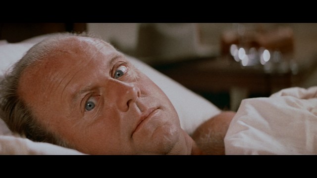 A close-up of a man with a concerned expression, with blurred figures in the background, set in a dimly lit interior.