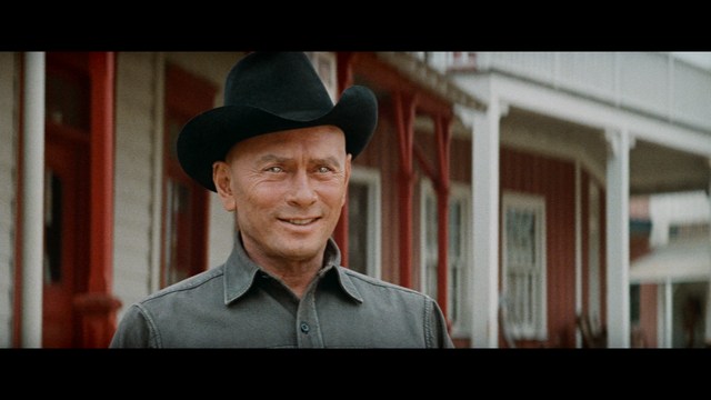 Close-up of a man with a serious expression, seated in a dimly lit area, with soft lighting from background lamps.