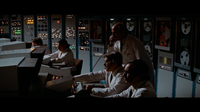 A close-up shot of a young man with short hair and a gray sweatshirt, looking intently at the camera, with a woman sleeping beside him in the background.