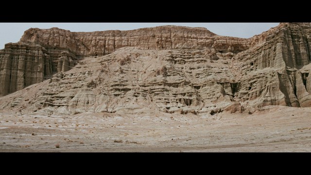 A woman seated at a restaurant table, thoughtfully resting her chin on her hand while reading a newspaper. Blurred figures of two people can be seen in the background.
