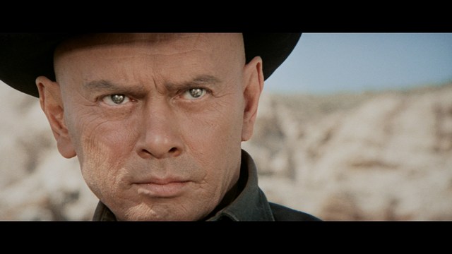 Two characters sitting at a table, one holding a small statue, with a surprised expression. The background features vibrant colors and patterns.