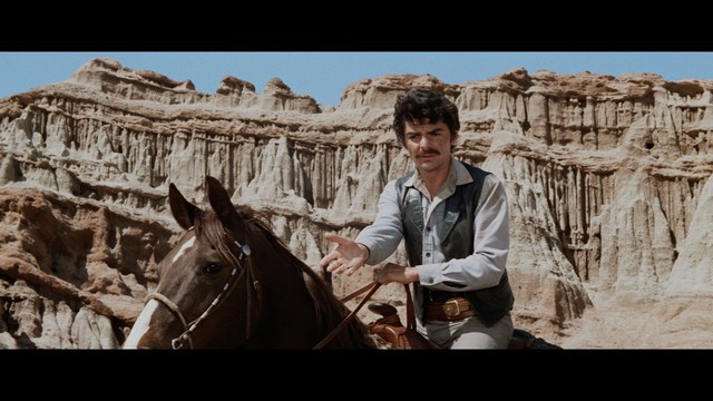 A serious-looking cardinal with a red hat and black robes sitting in a wooden chair in an office, surrounded by colorful posters and memorabilia.
