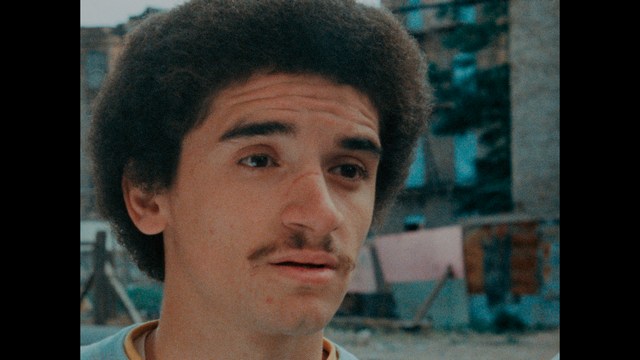 A man with a beard and mustache smiles warmly while standing on a boardwalk by the beach, with colorful flags overhead and a large building in the background.