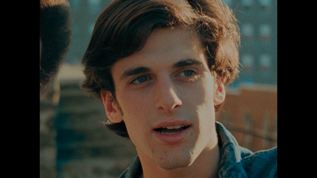 A serious-looking cardinal stands in front of a wooden backdrop with colorful posters related to Catholicism, holding a microphone at a press conference.