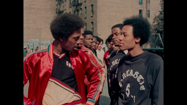 A woman with a pensive expression rests her chin on her hand, sitting at a table with kitchen appliances and items in the background.