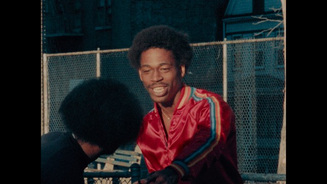 A man in a white suit and hat stands at a doorway, smiling, with a suburban landscape in the background.