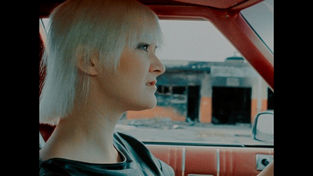 A man with large, feathered angel wings stands in a room, wearing a black suit and looking directly at the viewer.