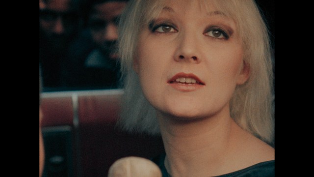 A woman with dark hair and a neutral expression sitting in a retro diner setting.