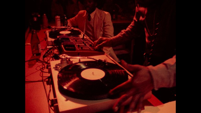 A scene featuring a man with short hair and a beard, wearing a brown jacket and purple shirt, standing outside a restaurant with bright yellow walls and a cartoon cow logo.