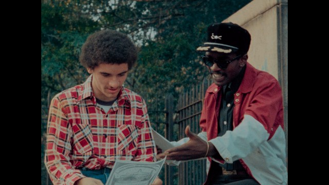 Close-up of a woman with long black hair sitting in a restaurant, looking concerned. A colorful painting is visible in the background, along with red bar stools.