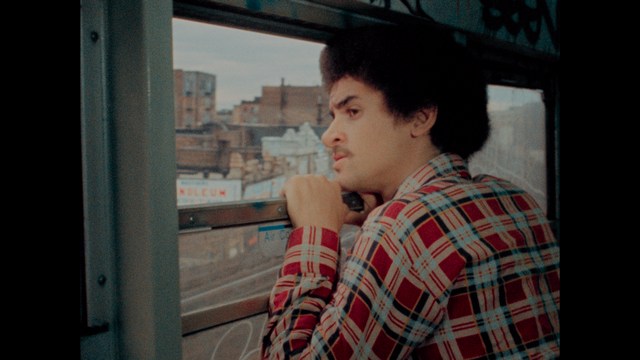 Two young men are looking at something off-camera, standing inside a building with bright lighting and seating areas visible in the background.