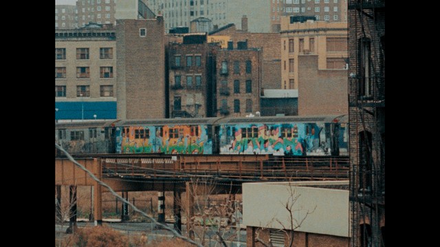 A close-up shot of a young man with short hair and a gray sweatshirt, looking intently at the camera, with a woman sleeping beside him in the background.