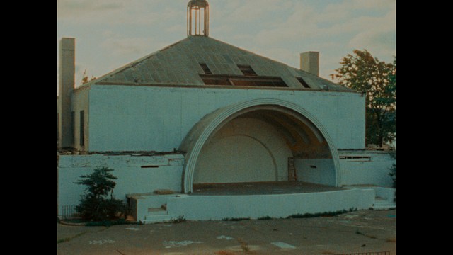 A woman seated at a restaurant table, thoughtfully resting her chin on her hand while reading a newspaper. Blurred figures of two people can be seen in the background.