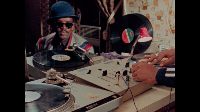 A man with dark, styled hair and a subtle smile seated at a restaurant table, with blurred diners in the background.
