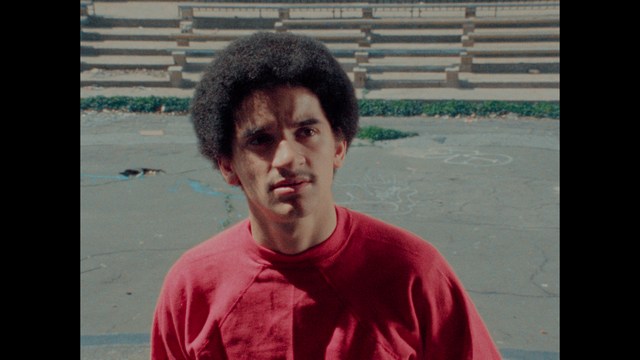 Two characters sitting at a table, one holding a small statue, with a surprised expression. The background features vibrant colors and patterns.