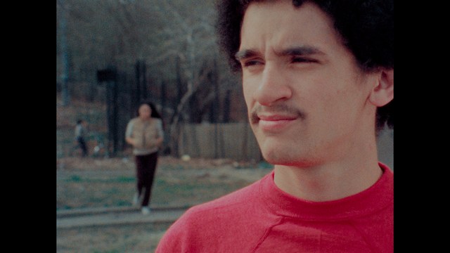 A serious-looking cardinal with a red hat and black robes sitting in a wooden chair in an office, surrounded by colorful posters and memorabilia.