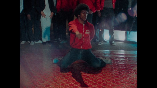 A scene featuring a man in a bishop's attire speaking at a podium surrounded by microphones, with a woman in a red dress beside him and a crowd of onlookers in the background.